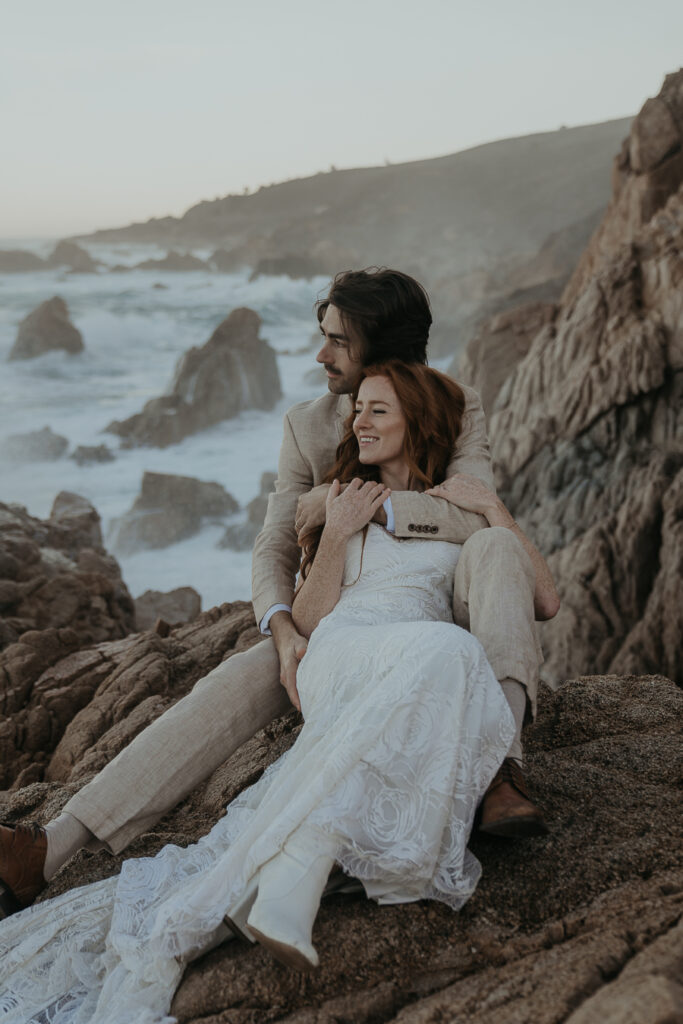 Bride and groom sitting on rocks holding each other overlooking the ocean in Big Sur