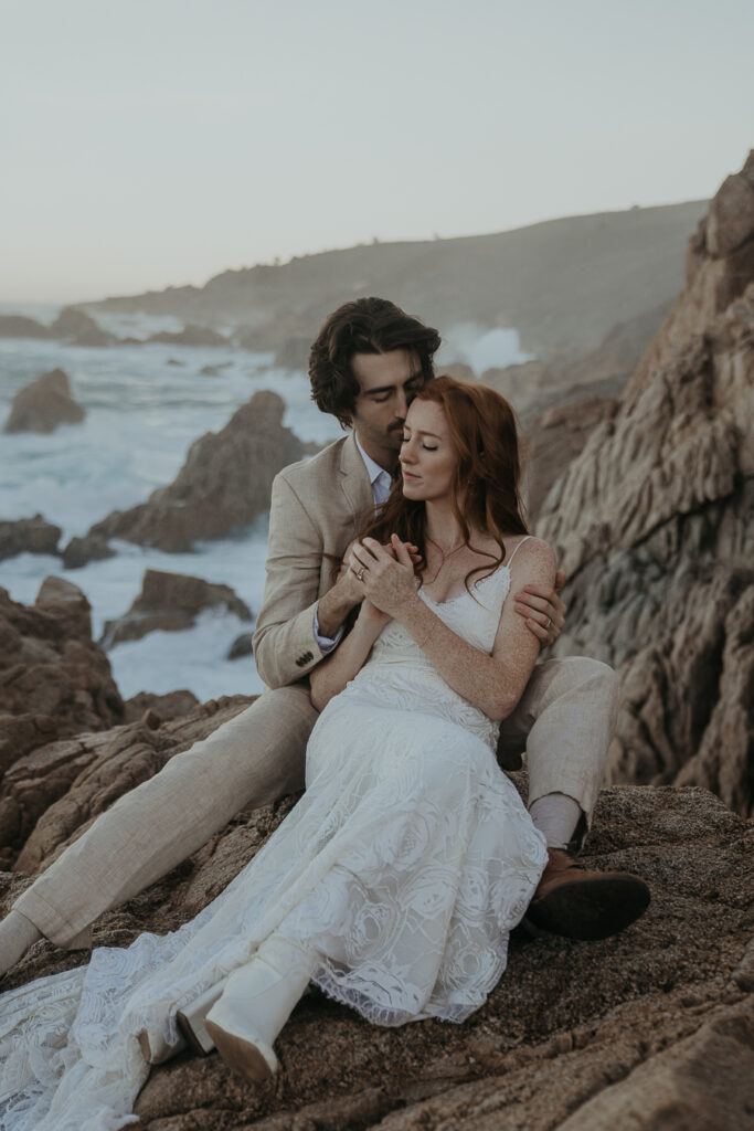 Bride and groom holding each other sitting on rocks overlooking the ocean