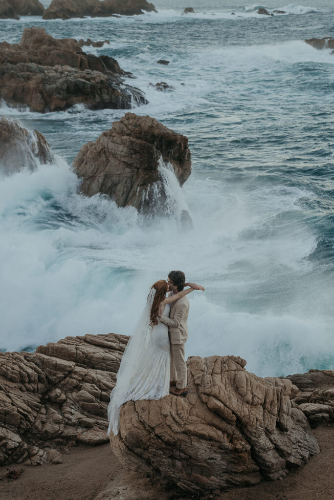 Bride wearing lace gown and long veil kisses groom wearing tan suit standing on rock in front of the ocean in Big Sur