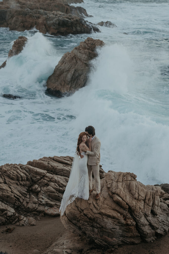Groom wearing tan suit kisses bride on the cheek while standing on rock in front of the ocean