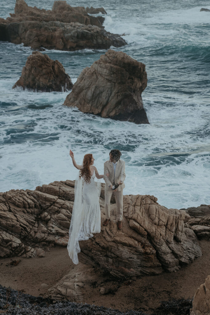 Bride wearing lace gown and long veil jumps up and down with arm raised while groom wearing tan suit pops champagne while standing on a rock with ocean in background