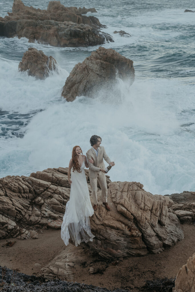 Bride and groom wearing wedding attire excitedly spray champagne in celebration of their elopement in Big Sur