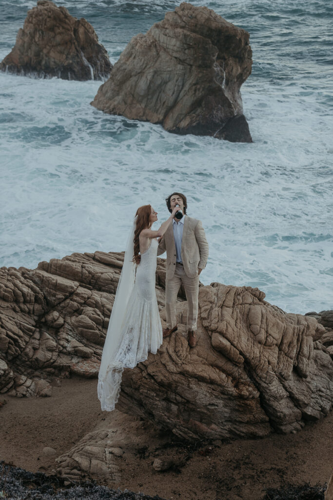 Bride wearing lace gown and long veil pours champagne into grooms mouth standing on a rock with ocean in the background