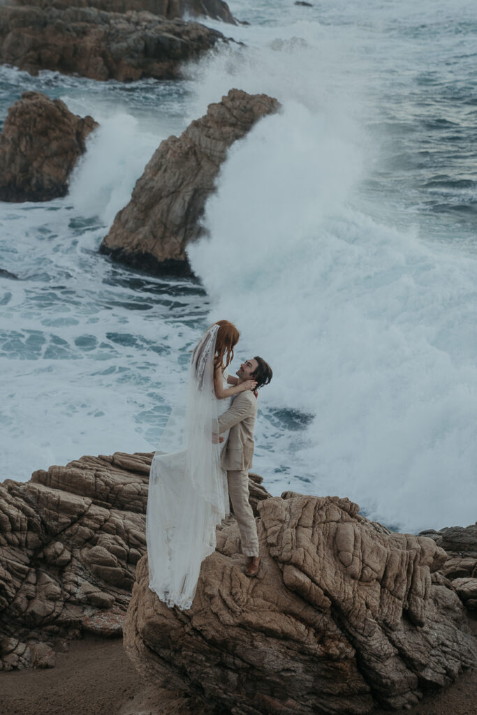 Groom wearing tan suit lifts bride wearing lace gown and long veil off the ground while standing on rock in front of the ocean in Big Sur