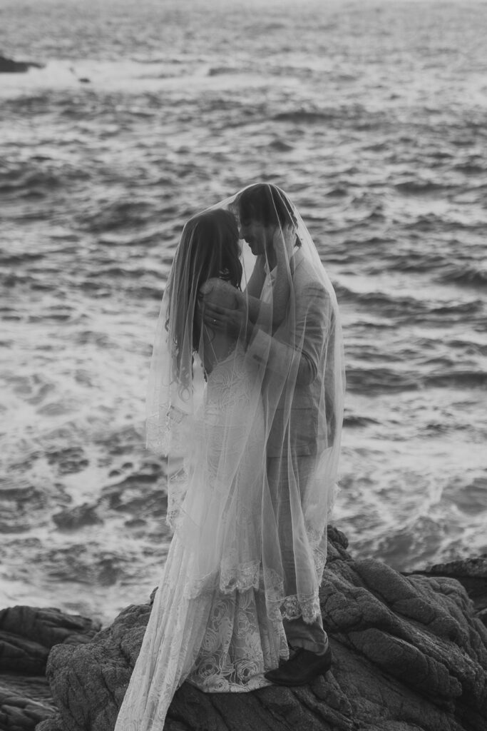 Black and white photo of bride and groom holding each other draped in brides veil while standing on rock above the ocean in Big Sur