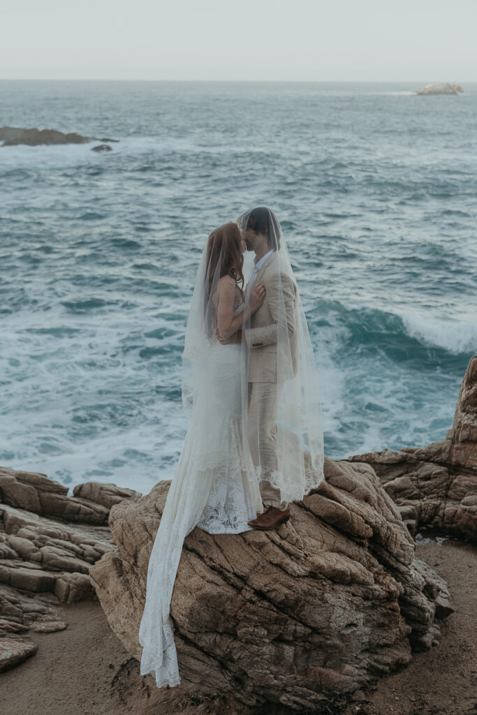 Bride and groom kissing while draped in brides veil standing on a rock in front of the ocean