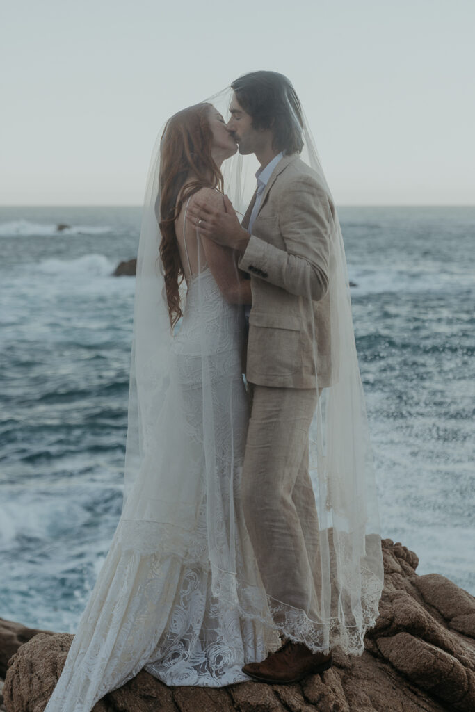 Bride and groom kissing while draped in a see through veil standing on rock above the ocean in Big Sur