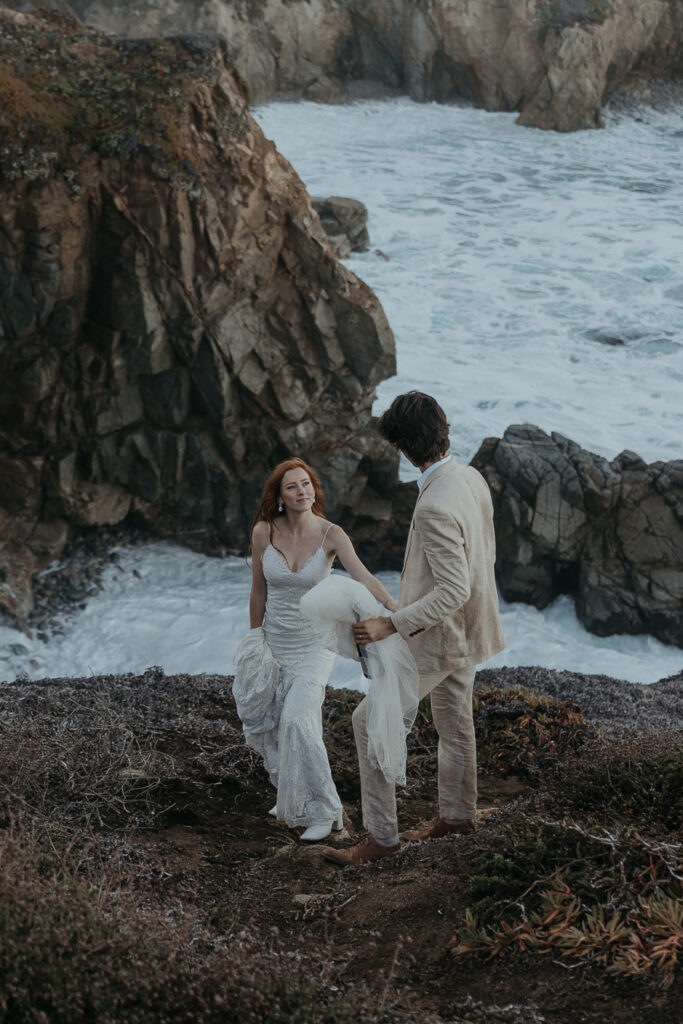 Bride and groom walking on a bluff trail above the ocean in Big Sur holding hands while groom holds brides veil and bottle of champagne in other hand