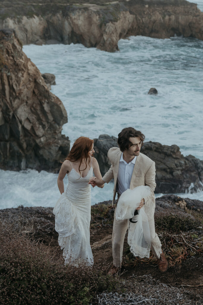 Groom leads bride down a trail on coastal bluff above the ocean in Big Sur while holding veil and bottle of champagne
