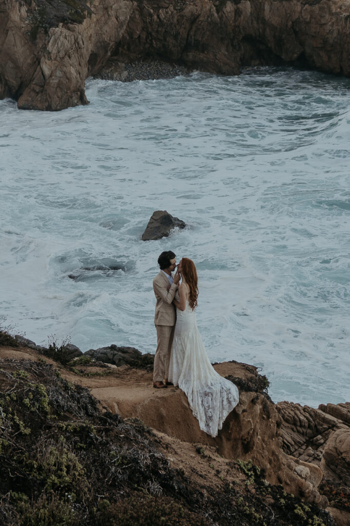 Bride wearing lace gown draped over rock leans in for kiss with groom wearing tan suit standing on a bluff overlooking the ocean in Big Sur