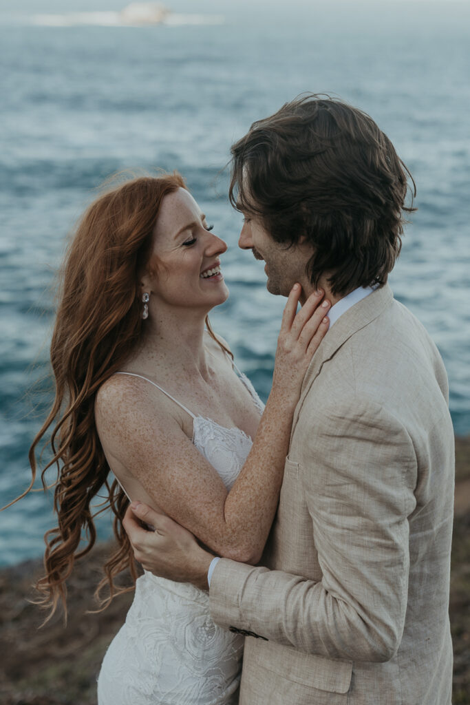 Close up photo of Bride and groom laugh while looking at each other in front of the ocean
