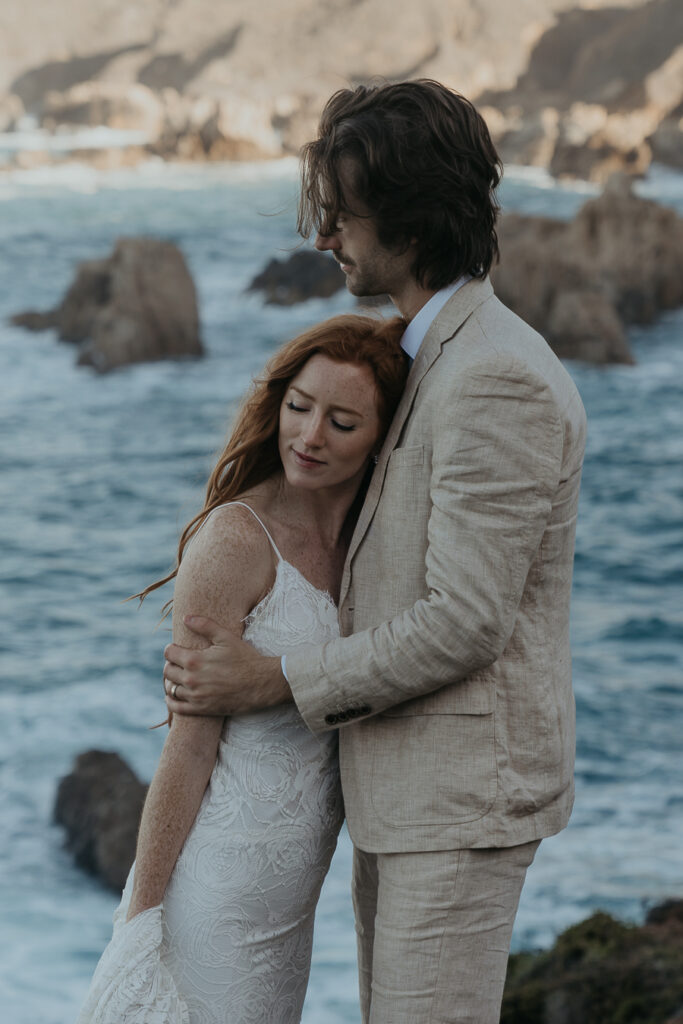 Close up photo of bride leaning head against grooms chest with rocky coastline and ocean in the background