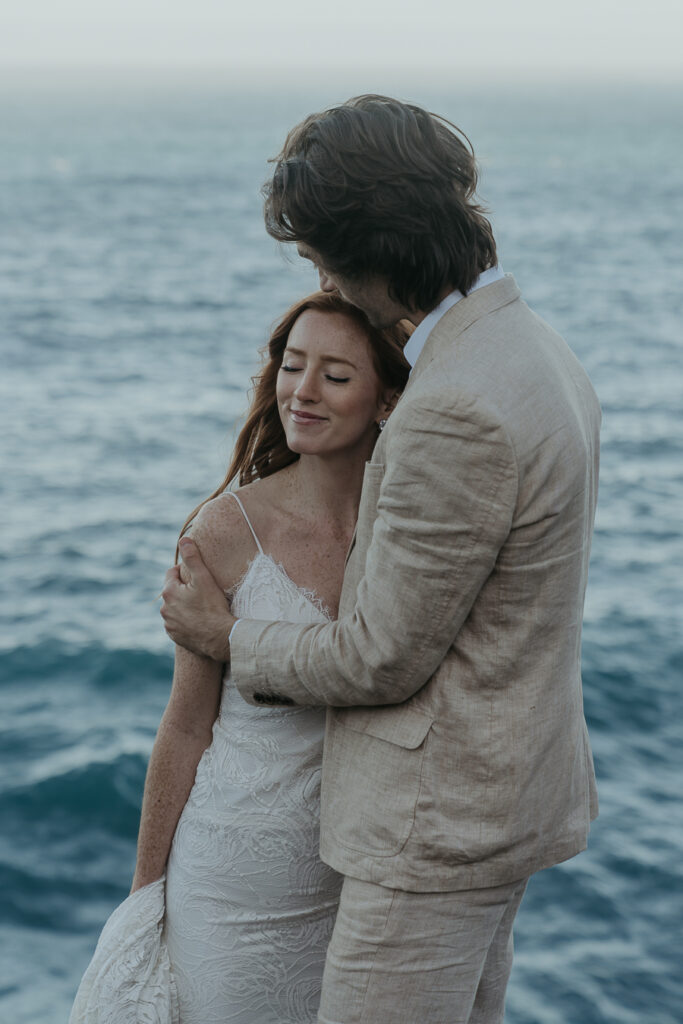 Groom wearing tan suit kisses top of brides head with ocean in the background