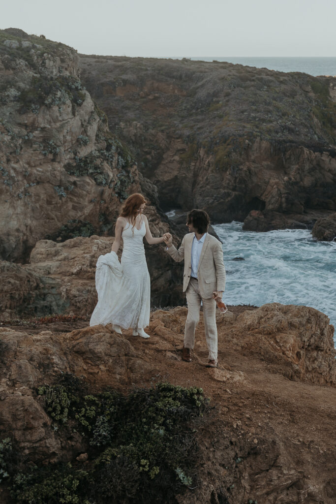 Groom wearing tan suit holds hand with bride wearing lace gown and white boots while walking down rocky trail by the ocean in Big Sur