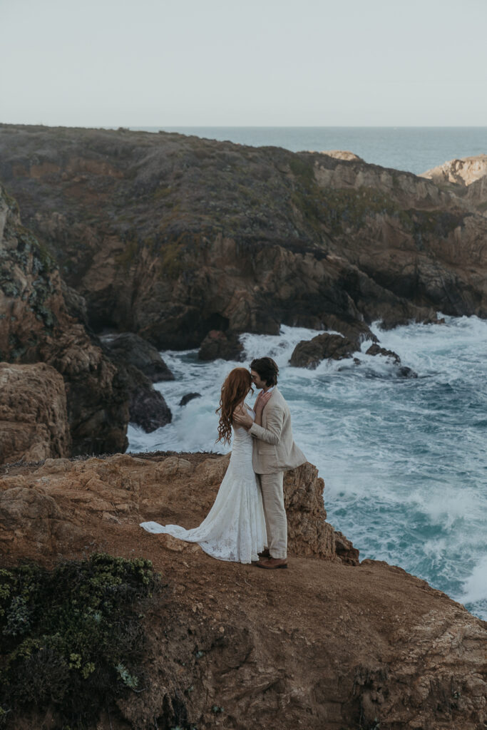 Bride and groom wearing wedding attire lean foreheads together standing on rocky cliff above the ocean