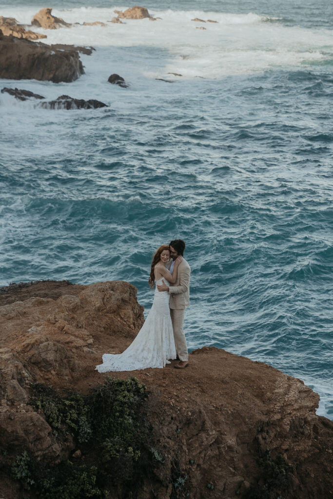Bride wearing lace gown and groom wearing tan suit hug smiling while standing on cliff above the ocean