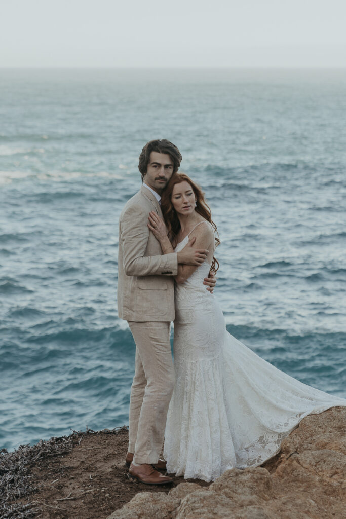 Bride and groom hold each other looking away standing on rocks in front of the ocean