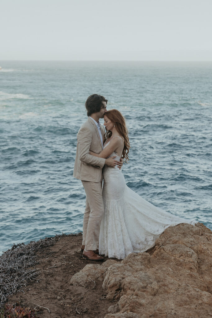 Bride wearing lace gown and groom wearing tan suit hold each other while groom kisses brides forehead with ocean in the background