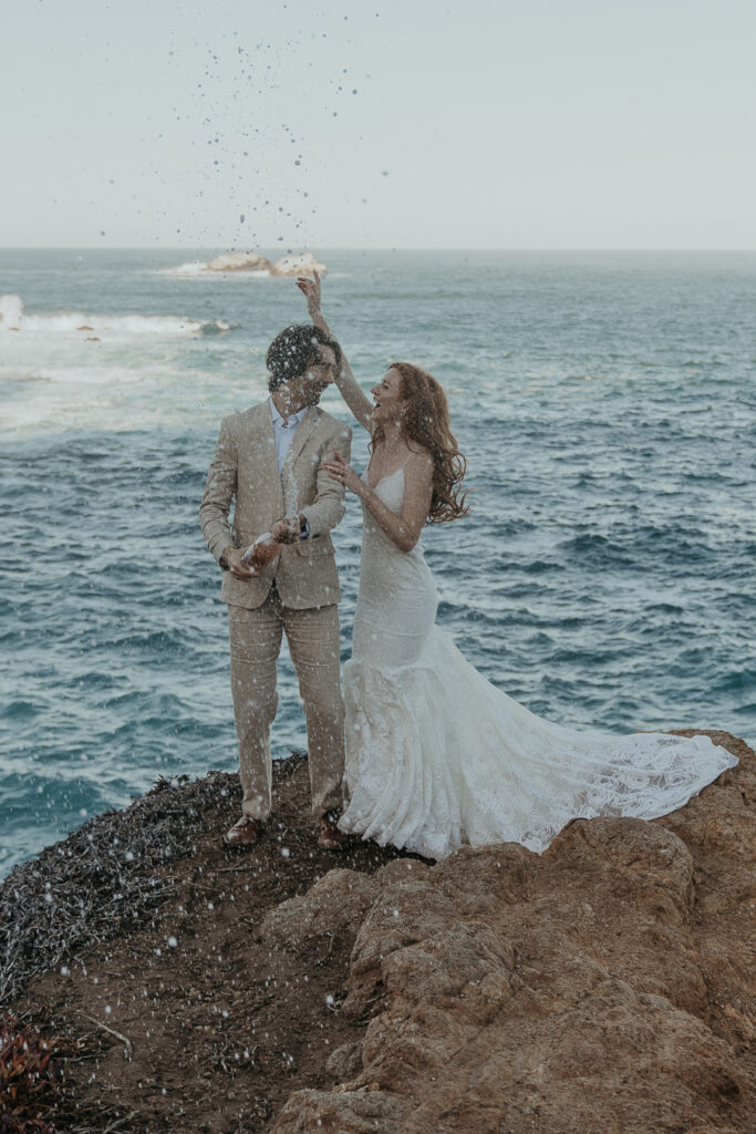 Bride wearing lace gown and groom wearing tan suit get excited while spraying champagne on a rock with the ocean in background