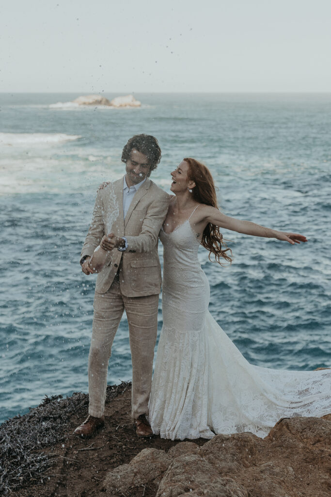 Bride wearing lace gown excitedly jumps up and down waving her arms while groom wearing tan suit pops champagne to celebrate their elopement in Big Sur