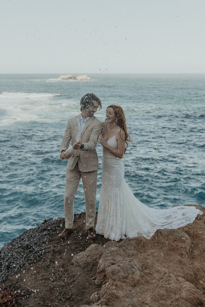 Bride wearing lace gown holds onto grooms arm while groom pops and sprays champagne to celebrate their elopement in Big Sur