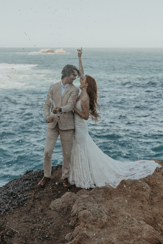 Bride wearing lace gown and groom wearing tan suit excitedly pop champagne to celebrate their elopement by the ocean in Big Sur