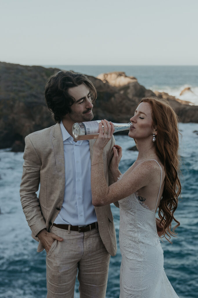 Bride wearing lace gown drinks champagne while groom wearing tan suit smiles at her with ocean in the background