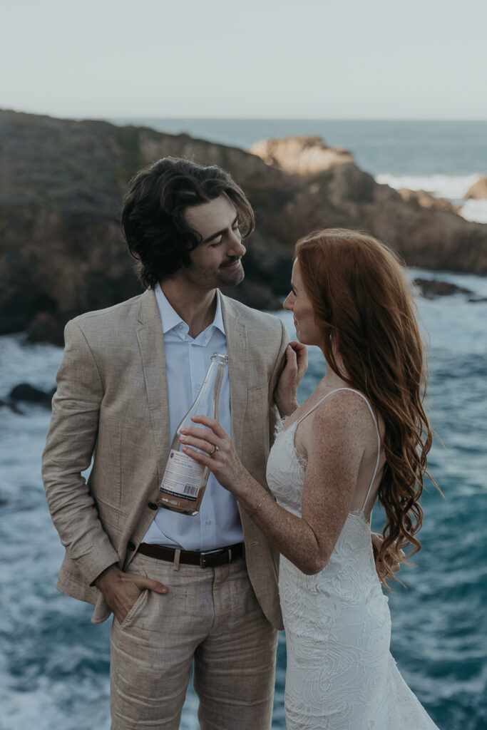 Bride wearing lace gown and groom wearing tan suit smile at each other while bride holds bottle of champagne standing in front of the ocean