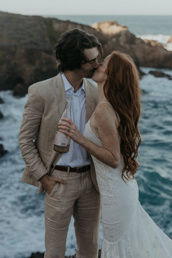 Bride wearing lace gown and groom wearing tan suit kiss while holding champagne with ocean in the background