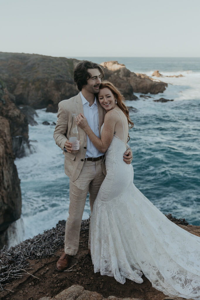Bride wearing lace gown and groom wearing tan suit hug each other while standing on bluff above the ocean holding champagne bottle
