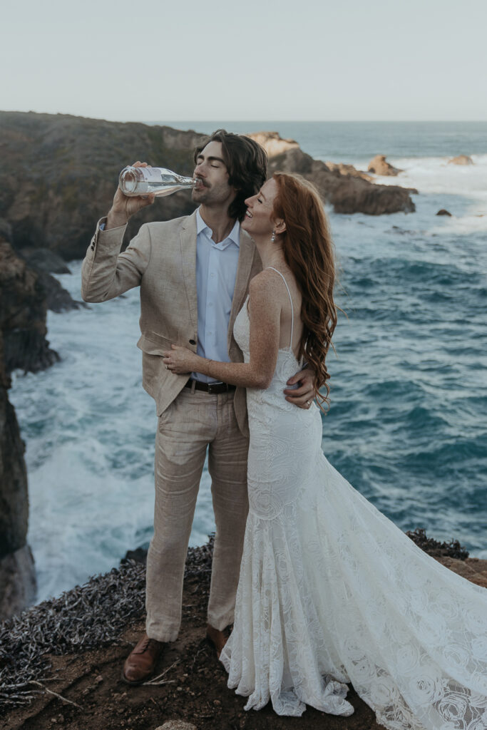 Bride wearing lace gown smiles at groom wearing tan suit while groom drinks from champagne bottle with arm around bride's back standing on coastal bluff over the ocean