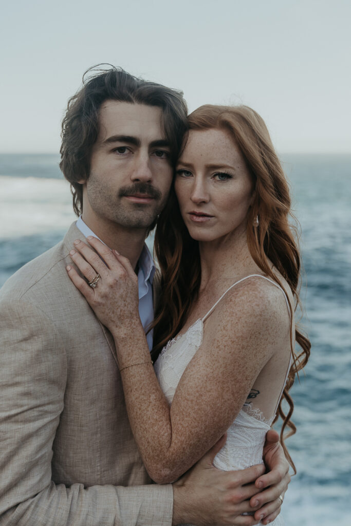 Close up photo of bride and groom side by side looking directly into the camera with ocean in the background