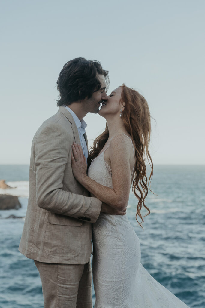 Bride wearing lace gown and groom wearing tan suit go in for a kiss while smiling standing with ocean in the background