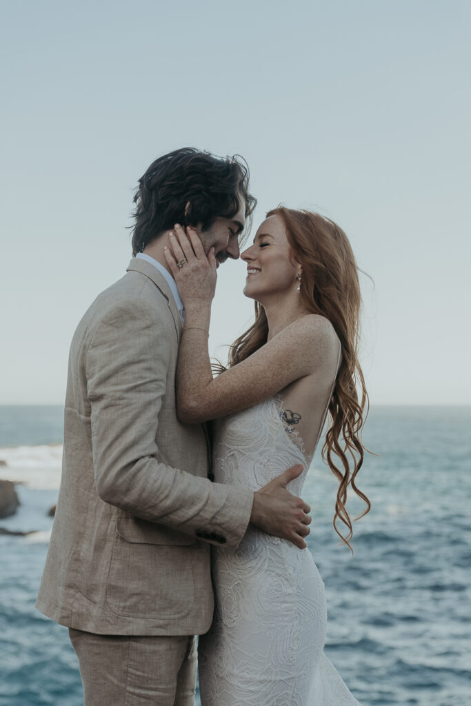 Bride wearing lace gown holds grooms face while groom wearing tan suit holds brides lower back laughing and smiling at each other with ocean in the background after big sur elopement