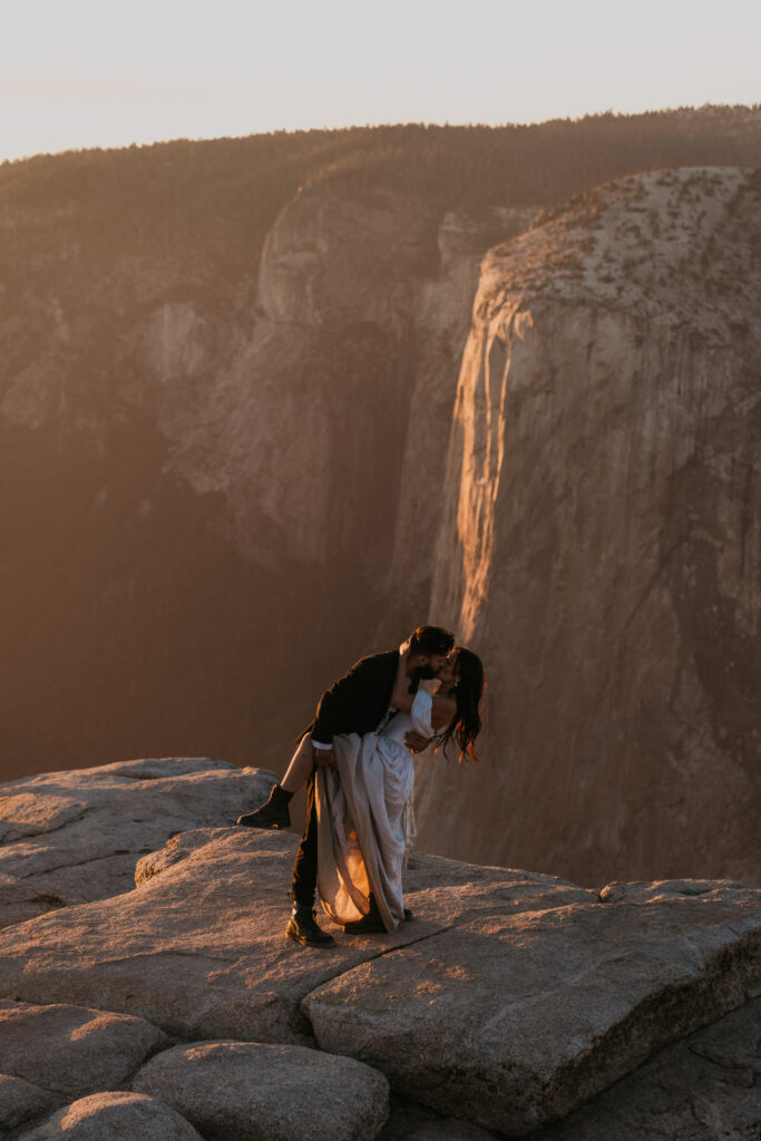 Groom dips bride for a kiss standing on a cliff in Yosemite with El Capitan in the background lit up by the sunset