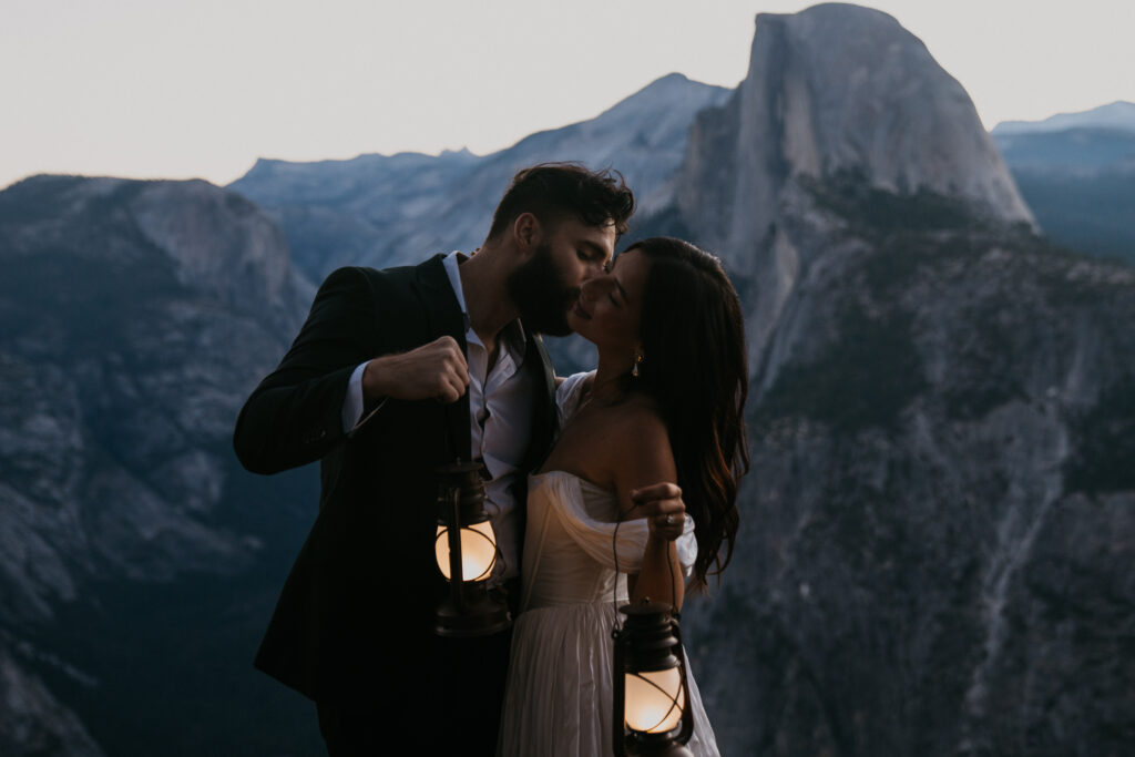 Groom kisses bride on the cheek while holding lanterns at sunrise in front of Half Dome