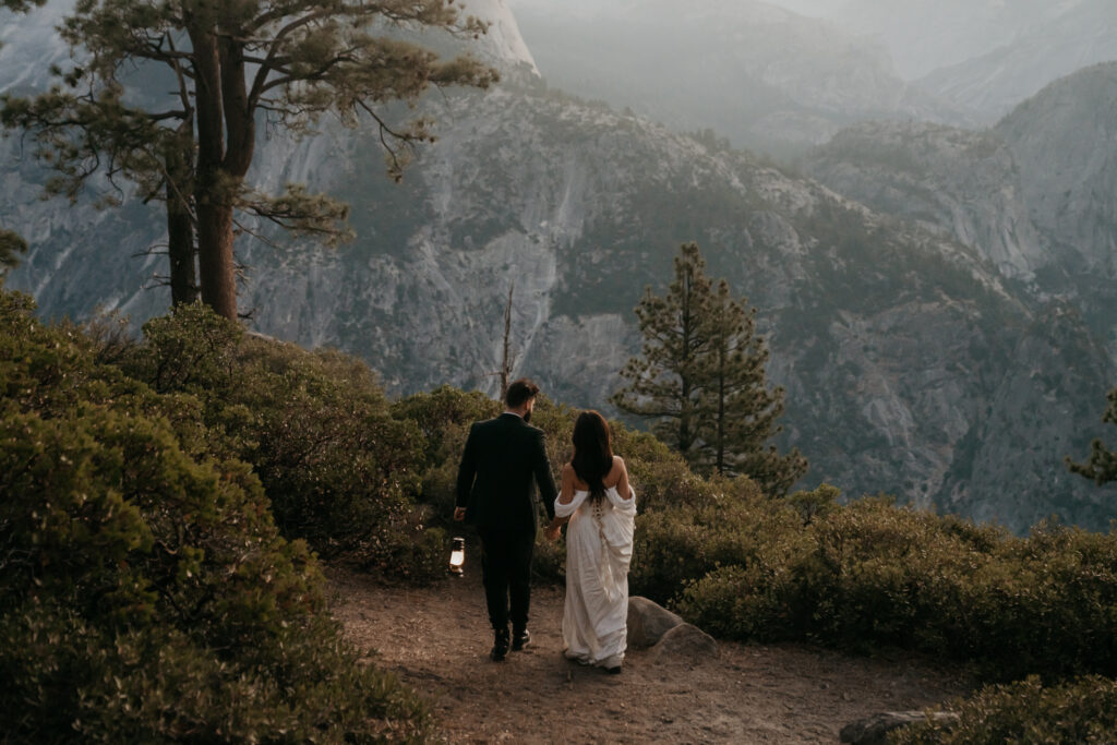 bride and groom walk away from camera hand in hand holding lanterns at Glacier Point surrounded by foliage and trees with the Sierras in the background